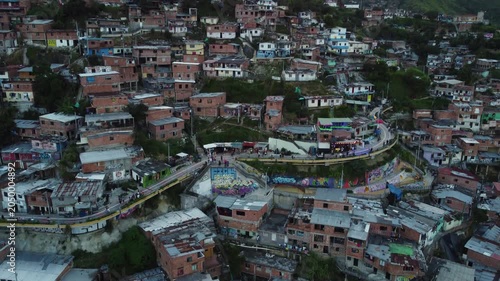 The pedestrian walkway at the top of Comuna 13, Medellin, Colombia, full of murals as well as shops catering to visitors, aerial ascending evening view of built-up hillside