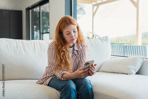 Obraz na płótnie Young woman with red hair sitting comfortably on a white sofa, engrossed in usin