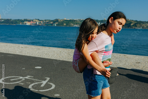 Older Sister Carrying Younger Sister Along a Waterfront Promenade