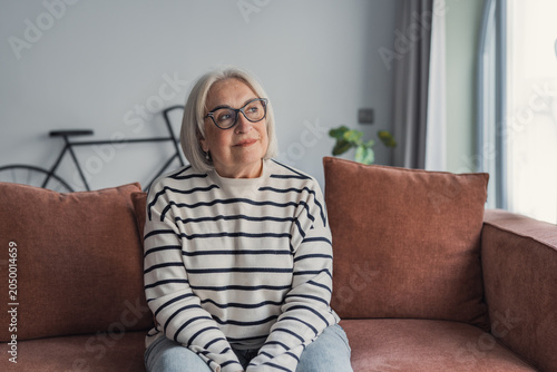 Foto Elderly woman with gray hair and glasses sitting on a brown sofa in a bright liv