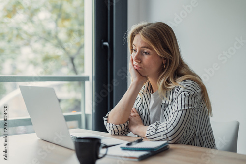 Canvastavla Young woman sitting at a desk with a laptop, looking away with a bored and tired