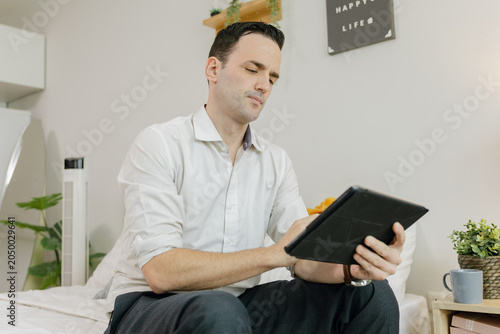 Photos A man sitting on a bed uses a tablet in a modern, cozy room with plants and decor in the background