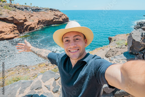 Young man wearing a straw hat and smiling at the camera, capturing a joyful seaside self-portrait with outstretched arms highlighting the beautiful rocky coastline and blue ocean