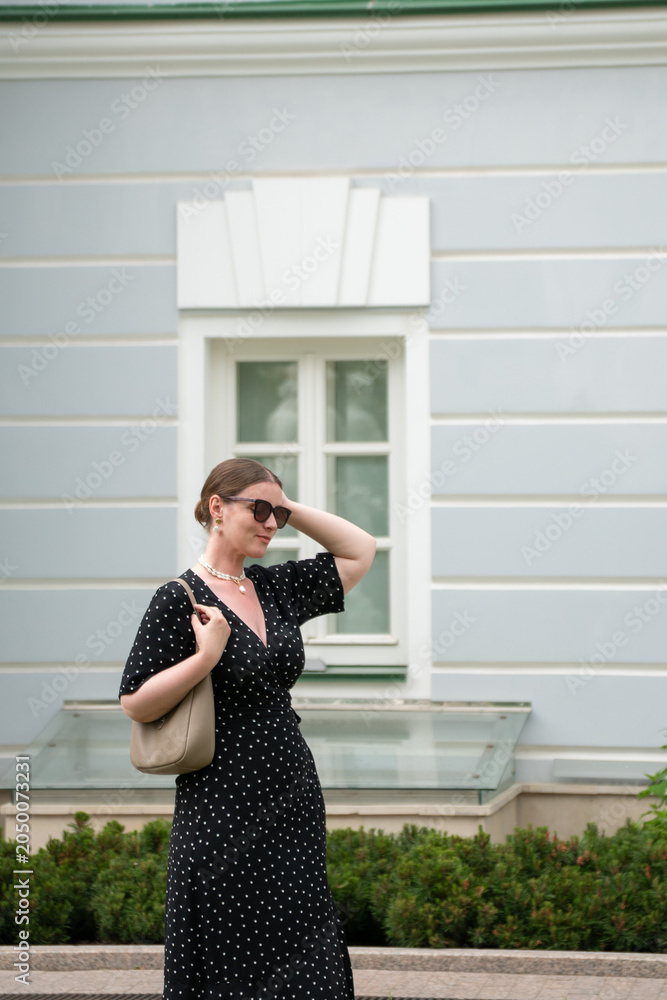 Fototapeta premium Woman in black polka dot dress, minimalist historic facade and landscaped surroundings creating elegant urban lifestyle scene. Cultural tourism, lifestyle, heritage, urban travel