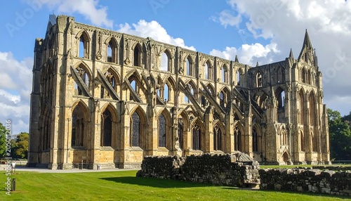 Impressive stone ruins of a large architectural structure, bathed in sunlight. Expansive grassy area surrounds the remnants. Sky shows fluffy clouds