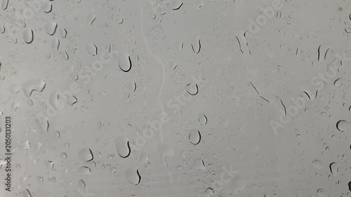 Close-up of water droplets and rain streaks on a clear glass window surface against a gray, overcast background.