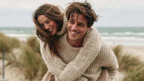 Happy young couple embraces on windy beach in knitted sweaters, smiling together near dunes and ocean.