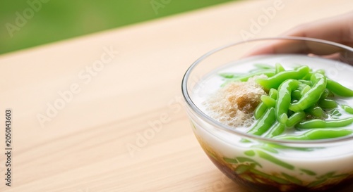 A hand holds a glass bowl of Sayur Asem on a wooden table outdoors.