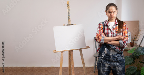 фотографія Upset female artist painter working in art studio with blank canvas looking displeased, standing with arms crossed
