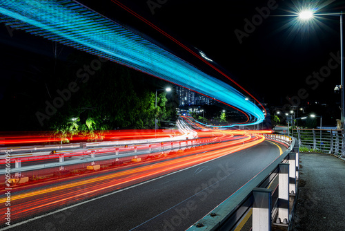 Canvastavla Long exposure shot capturing colorful light trails from vehicles on a curved road at night, with glowing cyan LED structure overhead and city lights in the background, South Korea