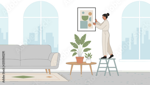 Young woman standing on stepladder to hang framed abstract art piece on wall in modern bright living room with large windows.
