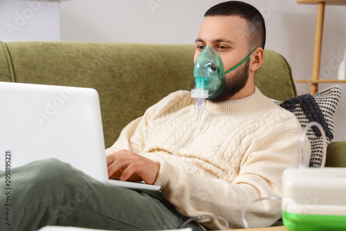 Young man with laptop using nebulizer on sofa at home