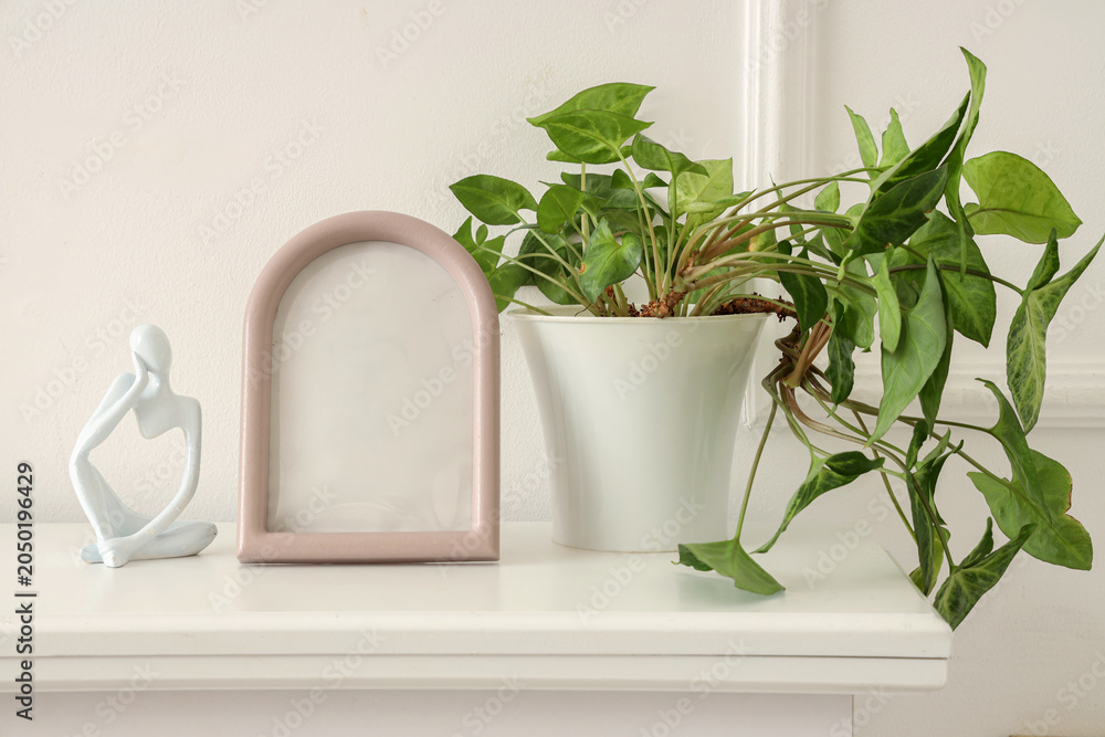 Obraz premium Blank frame with plant and statuette on shelf near light wall