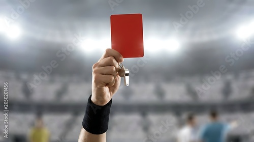 Referee hand holding red card and whistle for expulsion of player during soccer game on stadium field dramatic lighting bright.