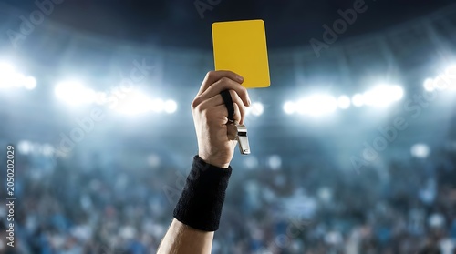 Referee hand raises yellow card and blows whistle in stadium filled with cheering fans under bright stadium lights.