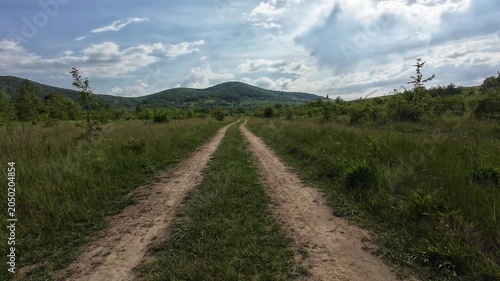A vehicle drives along a scenic dirt road through a peaceful countryside landscape surrounded by lush green meadows, trees, and rolling hills beneath a dramatic cloudy sky