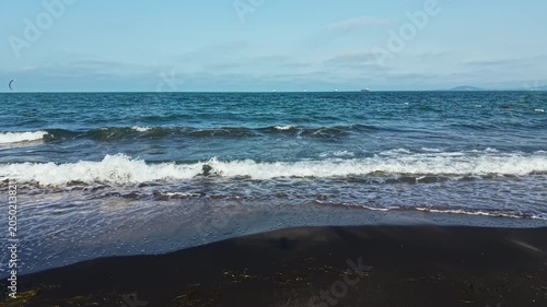 People enjoy activities at the beach while waves hit the sand on a clear day. Kitesurfers ride the water, and ships sail in the distance under a blue sky.