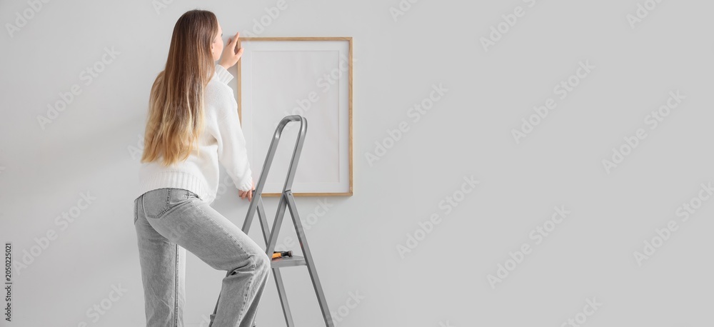 Εκτύπωση καμβά Woman on stepladder hanging blank picture frame on wall in modern minimalist hom