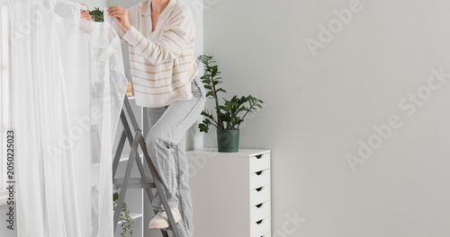 Tableau sur toile Young woman on stepladder hanging curtain in living room