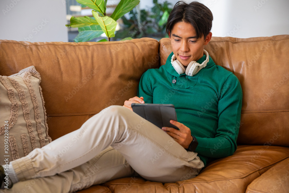 Fototapeta premium Asian man sitting on tan leather couch in living room holding dark tablet wearing headphones