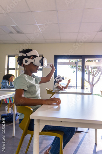 Classmates with African American male child sitting in yellow chair at white desk using VR headset