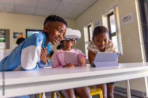 Three African American children gathering around white table at school using VR headset and tablet