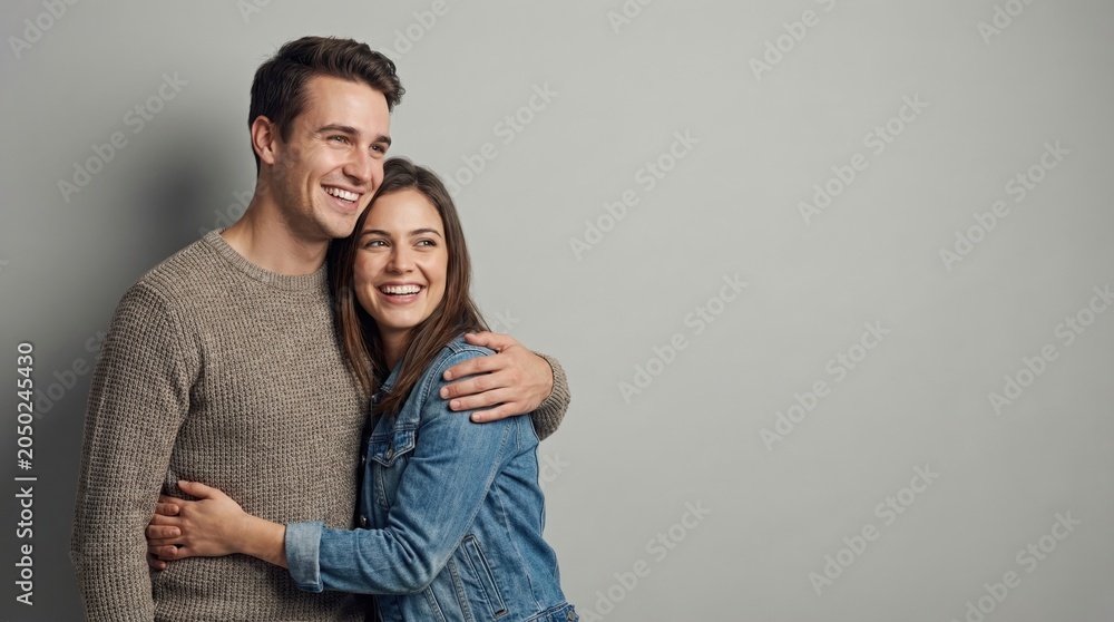 Fototapeta premium happy caucasian couple embracing and looking away against grey background. smiling man hugging woman in denim jacket. love, relationship concept. copyspace, banner, website header.