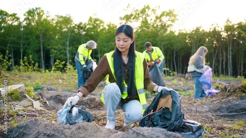 Asian female volunteer collecting waste during outdoor cleanup. Woman holding trash bag and removing litter from ground. Child and adults helping with cleanup work nearby. Eco activity in nature.