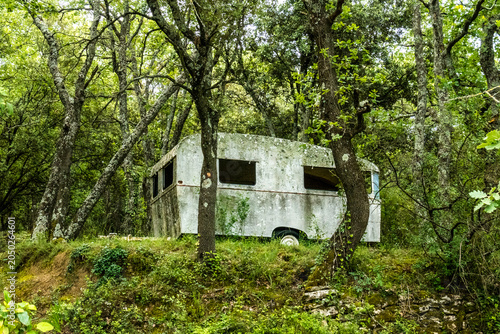 An abandoned, overgrown caravan sits hidden among dense green woodland trees, slowly being reclaimed by nature in a forest setting, near Castillon du Gard, France