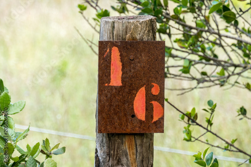 A rusty metal number 16 sign with orange stencilled numerals attached to a weathered wooden post amid green foliage, Castillon du Gard, France