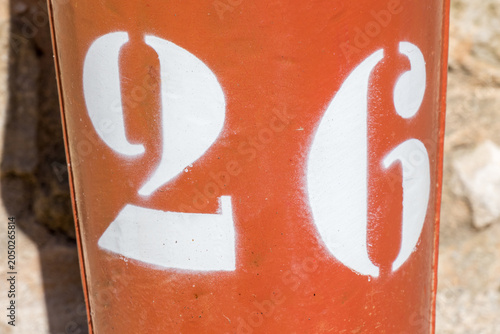 Bold white stencilled number 26 on a bright orange painted post or pillar against a rough stone wall in a French village