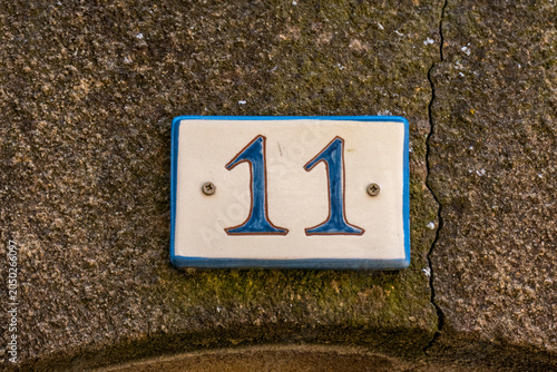A ceramic house number 11 plaque with blue numerals on a cream tile, mounted on a weathered stone wall in a French village