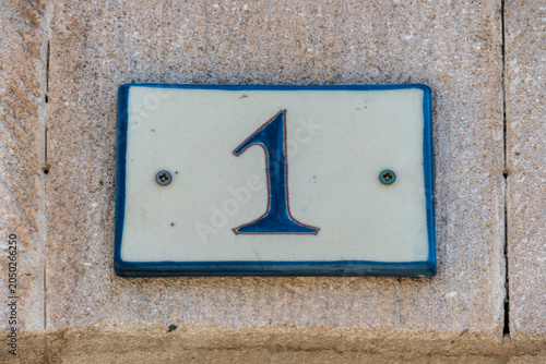 A ceramic house number 1 plaque with blue numerals on a cream tile, mounted on a weathered stone wall in a French village