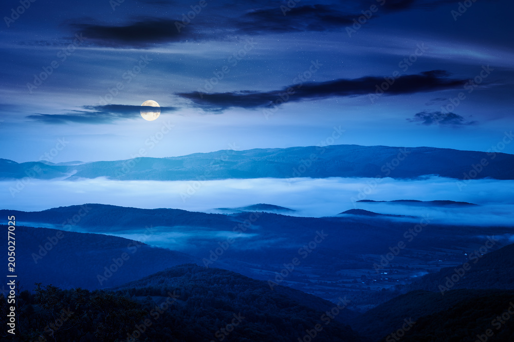 Fototapeta premium foggy carpathian countryside scenery in summer at night. mysterious view of mountainous landscape with rural fields on rolling hills. full moon over fog in the valley. clouds above the mountain
