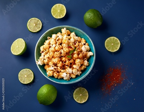bowl of popcorn with chili and lime seasoning in a teal bowl centered on a dark blue surface with lime wedge and green lime scattered around