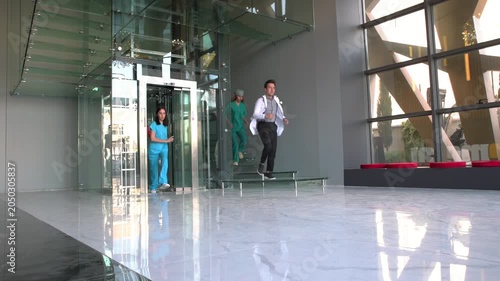 Doctors and nurses in scrubs and lab coats hurry through a modern hospital lobby with sleek glass walls and polished marble floors, indicating a busy medical facility