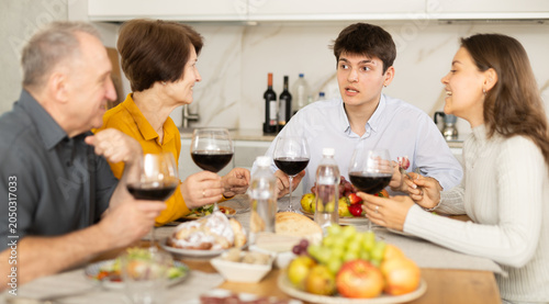 Fotografie Smiling young guy engaging in lively conversation with sister and elderly parents at family table with wine and fruits in cozy home kitchen