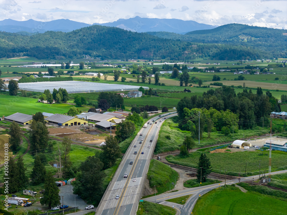 Fototapeta premium Aerial View Of Highway Cutting Through Farmland Valley With Mountains Near Abbotsford Mission BC, Canada