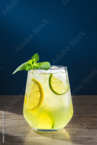 Refreshing cold lemonade with mint and lime slices. A misted glass with droplets stands on a wooden surface. The dark blue background highlights the bright colors. Photo