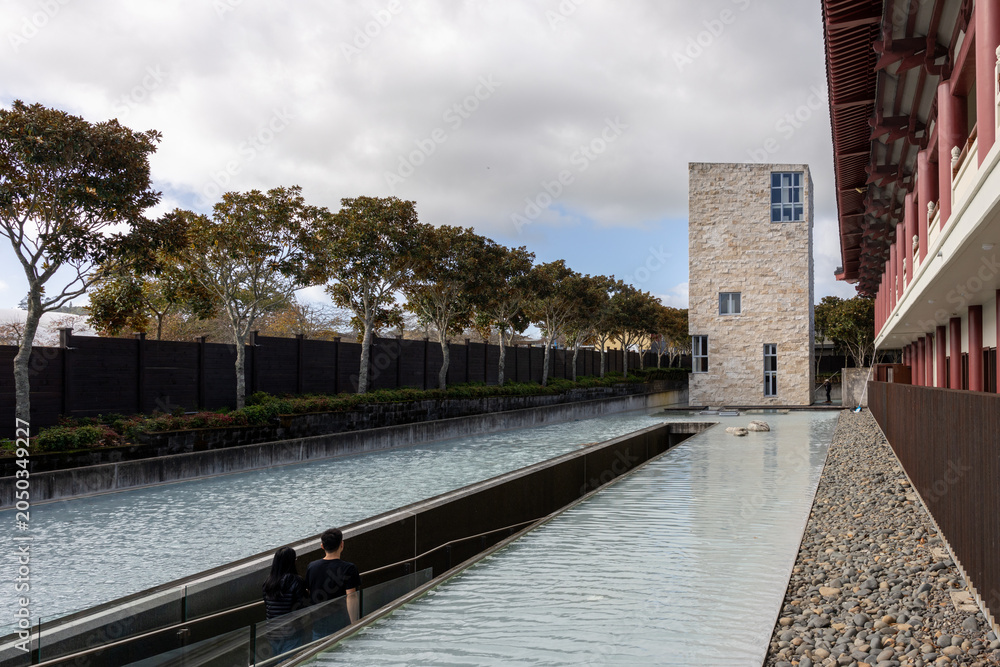 Fototapeta premium A couple walks by a tranquil water feature in front of a modern building with traditional architectural elements at Fo Guang Shan Buddhist Temple, Auckland, New Zealand.