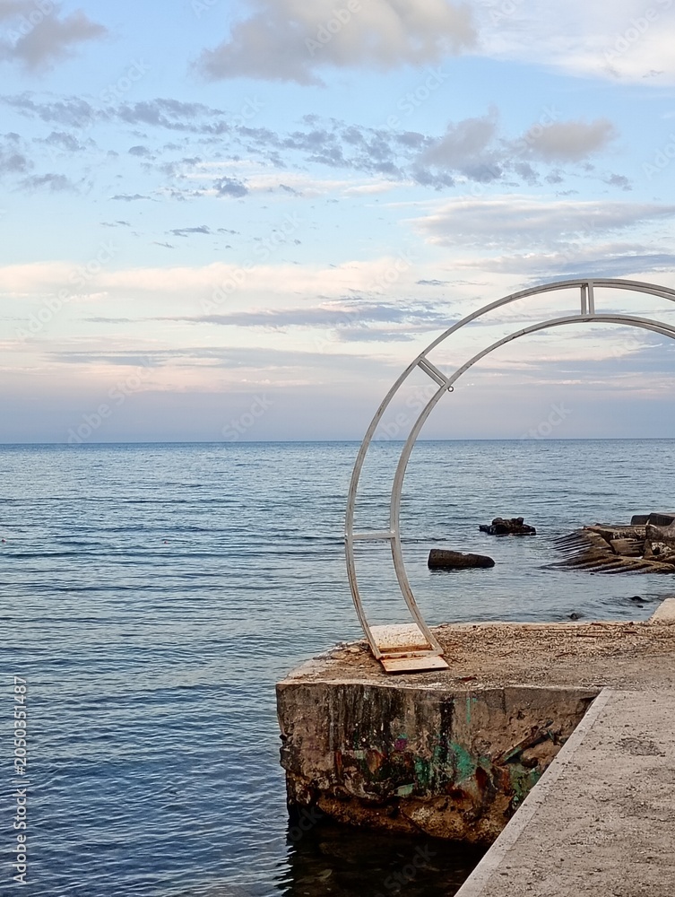 Obraz premium Minimalist metal wedding arch structure standing on a concrete seaside pier against a calm blue ocean horizon. High-quality seascape photograph perfect for romantic destination wedding planning websit
