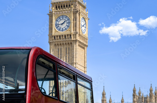 A close up of a traditional red double decker sightseeing tour bus with the historic Big Ben clock face and Elizabeth Tower rising behind under a blue sky in London England