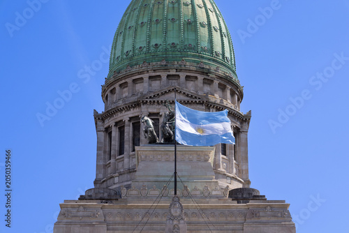 A close up view of the green copper dome and stone facade of the Palace of the Argentine National Congress featuring the national flag waving on a flagpole under a clear blue sky in Buenos Aires