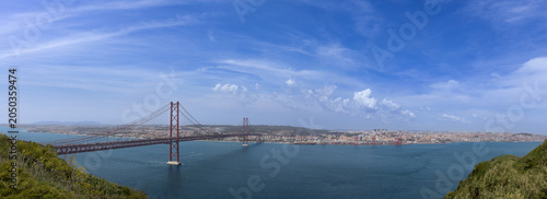 Panoramic view of the 25 de Abril suspension bridge spanning the Tagus river under a blue sky in Lisbon Portugal panorama