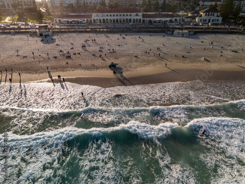 Bondi Beach aerial view crowded with swimmers, surfers, life guards and waves breaking in golden hour. Key buildings, pavilion and promenade can be seen in background.