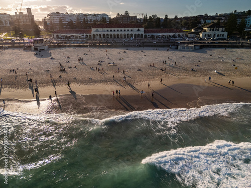 Bondi Beach aerial view crowded with swimmers, surfers and waves breaking in golden hour. Skyline, pavilion and promenade can be seen in background.