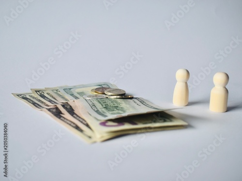 Photos Close-up of a stack of US dollar bills and metallic coins with two wooden peg doll figures standing in the background