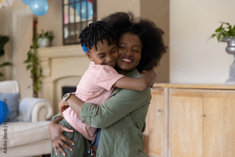 Obraz premium Mid adult African American mother and son hugging in living room with blue balloons, watch visible
