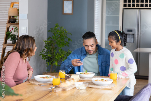Mid adult parents with child sharing meal around wooden dining table with bowls, orange juice, loaf
