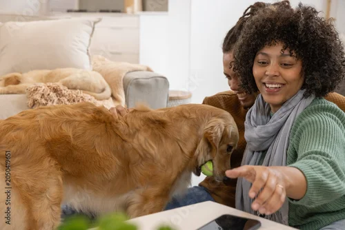 Fototapeta samoprzylepna Couple watching woman in green sweater reaching for phone at home, dog holding ball, copy space